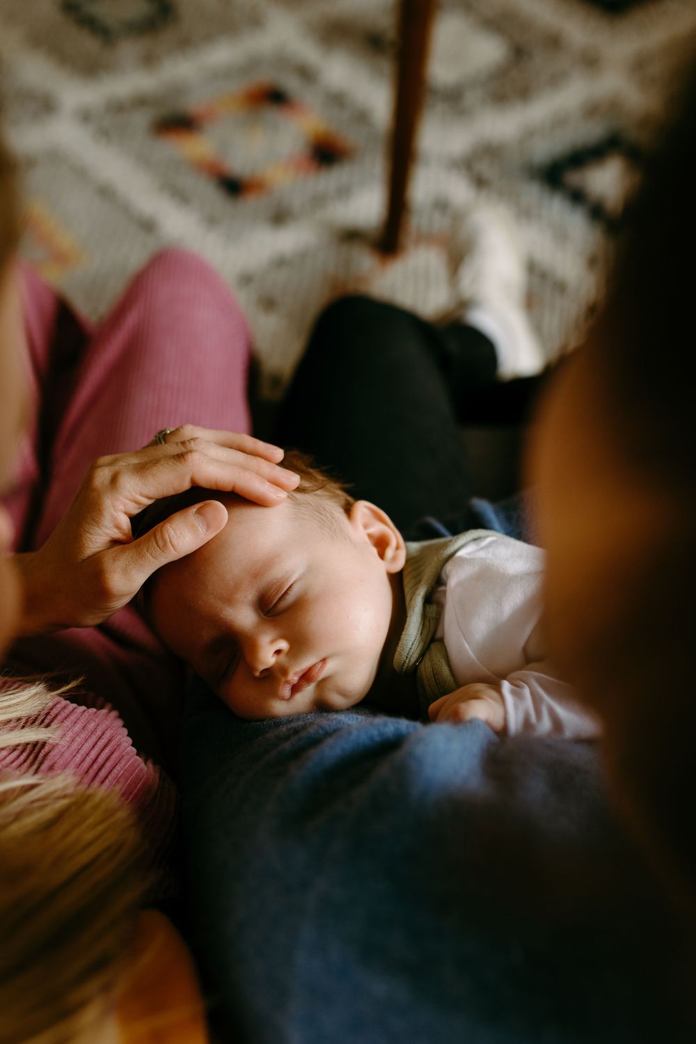 A sleeping newborn baby rests on a parent's lap while a gentle hand touches the infant's forehead.