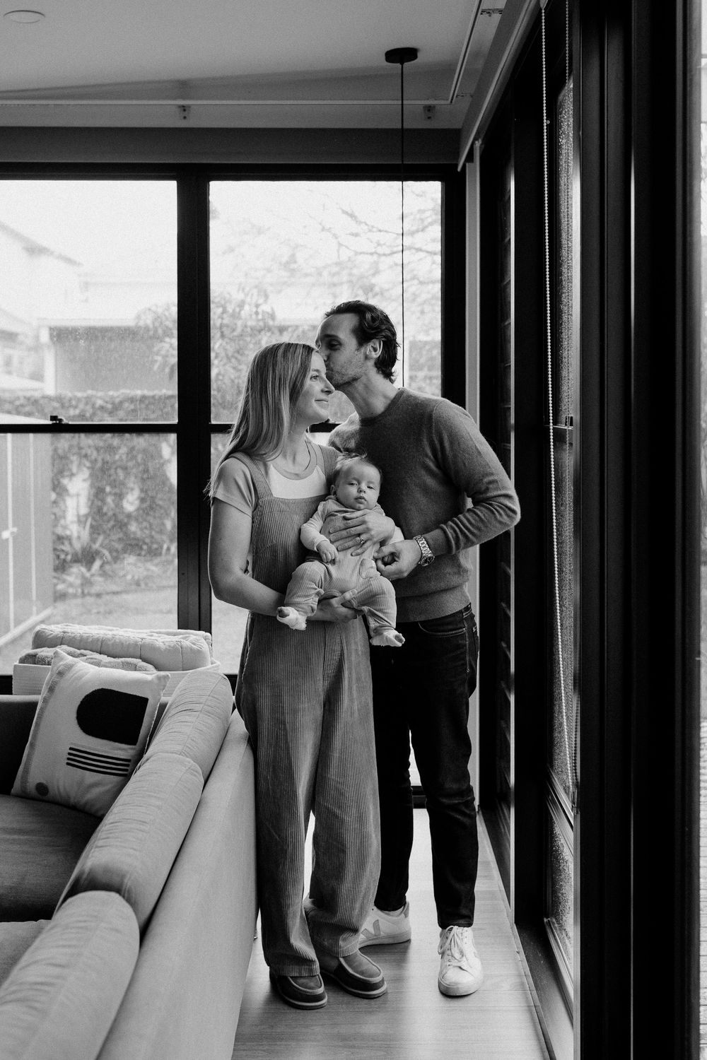 Black and white photo of a couple holding their newborn baby by large windows in a modern home.
