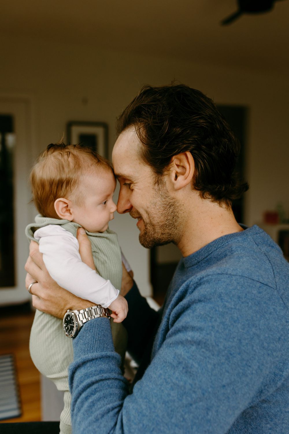 A smiling father tenderly touches foreheads with his baby while holding them indoors.
