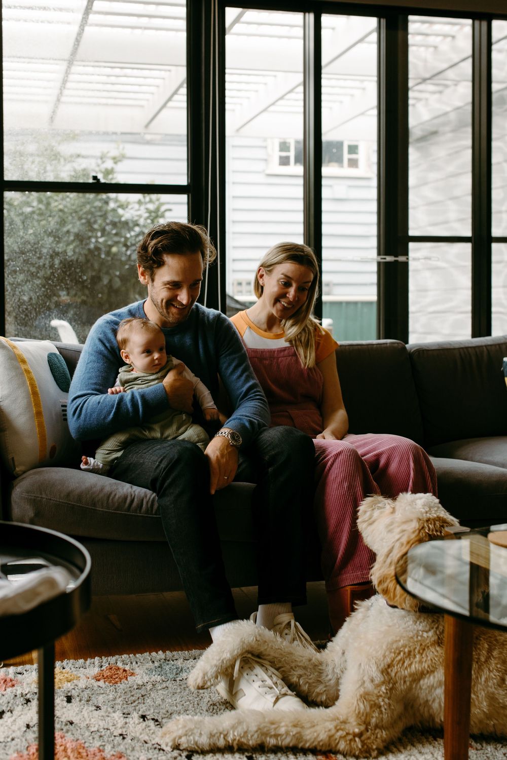 Happy family with baby and golden dog relaxing on gray sofa in modern living room with large windows.