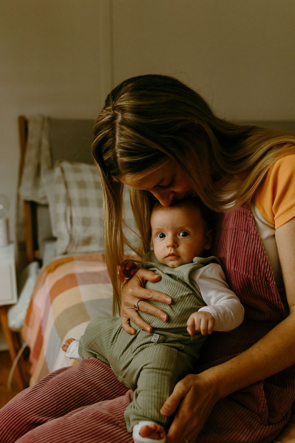 A mother tenderly kisses her alert baby dressed in green, sitting together in a warmly lit room.