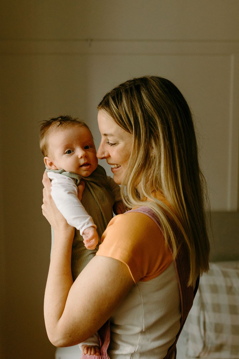 Smiling young mother in orange top gently holding and gazing at her newborn baby indoors.