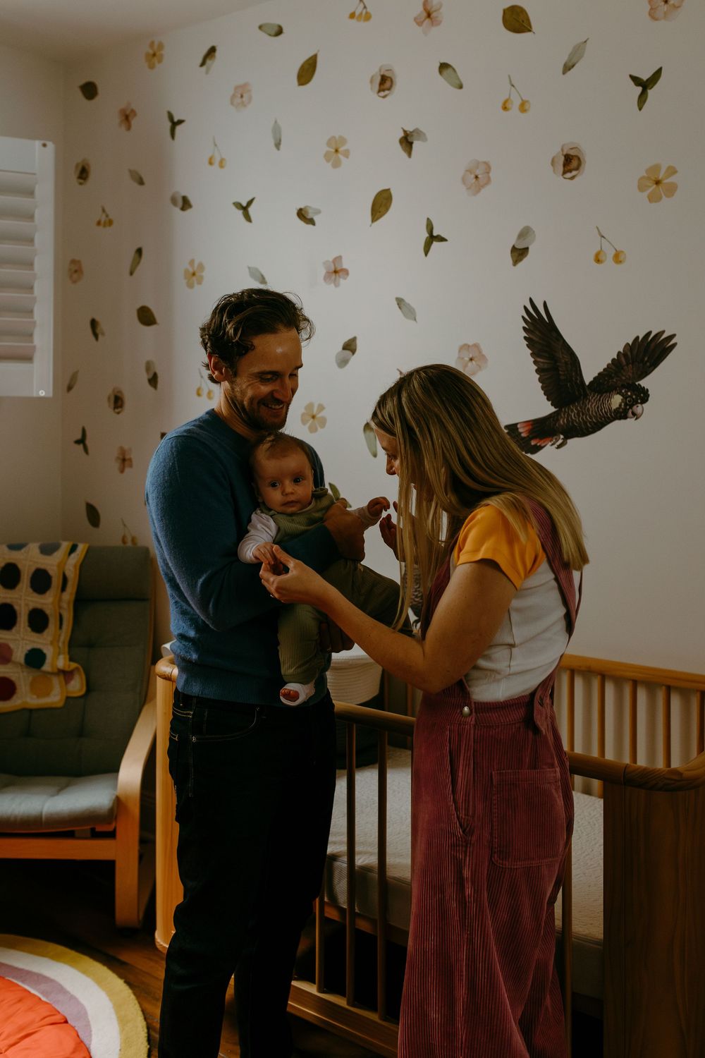 A couple admires their newborn baby in a nursery with floral wall decals and a wooden crib, as a parrot perches nearby.