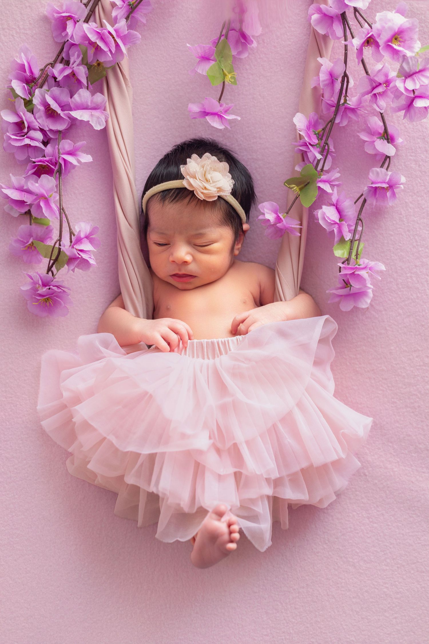 Sleeping newborn baby girl in pink tutu skirt and floral headband surrounded by purple cherry blossoms.