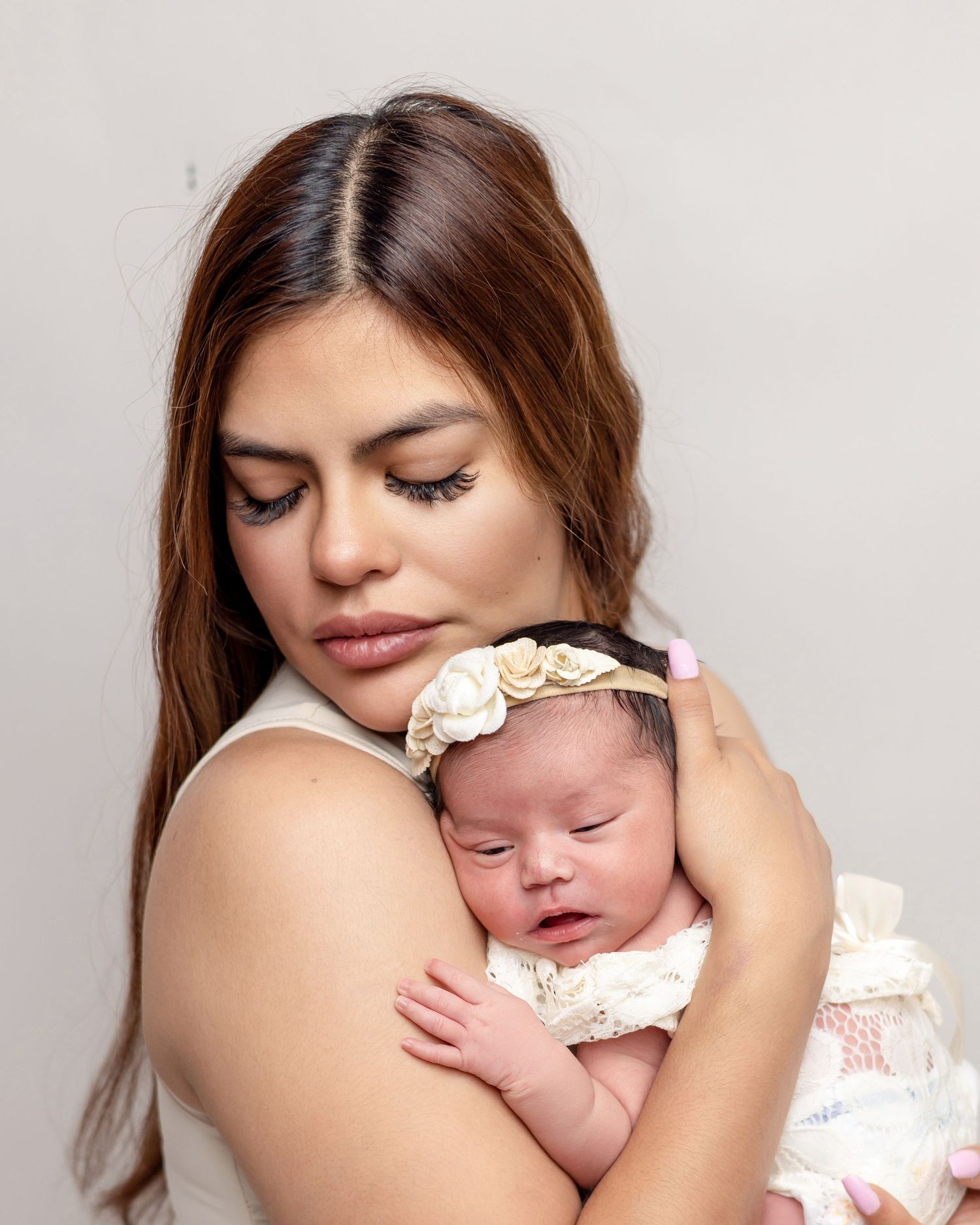 A young mother tenderly holds her sleeping newborn baby girl wearing a floral headband against her chest.