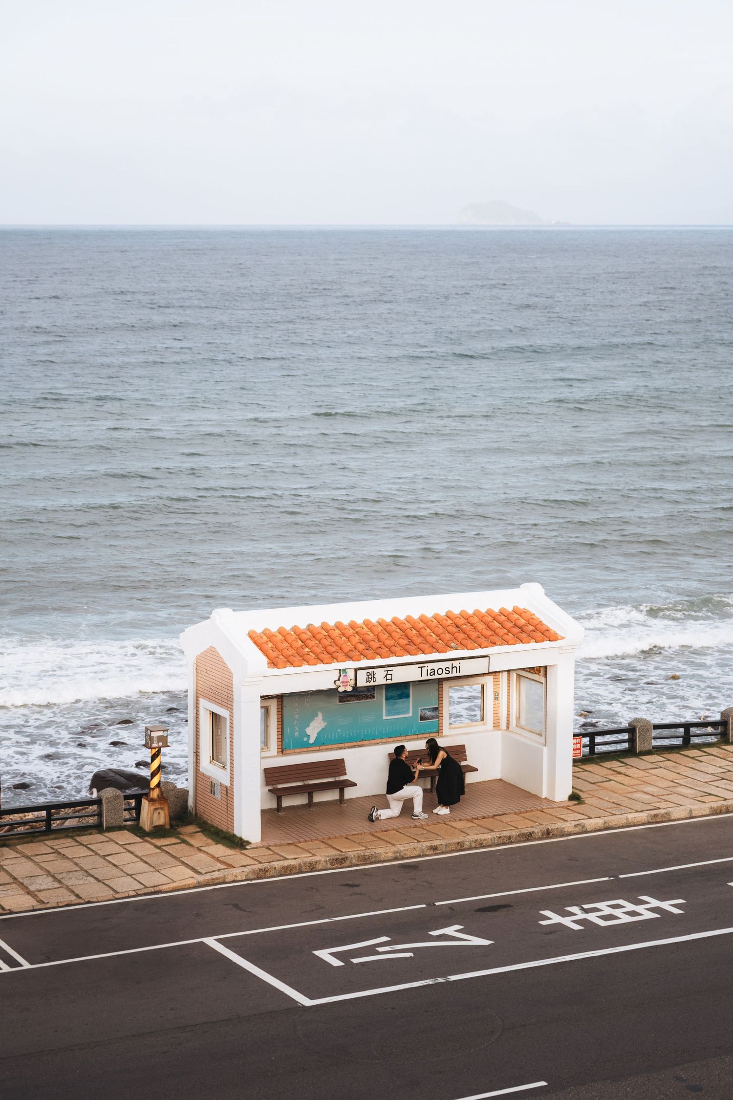 A white bus stop shelter with orange roof sits beside a coastal road in Japan, with two people waiting as ocean waves crash behind.
