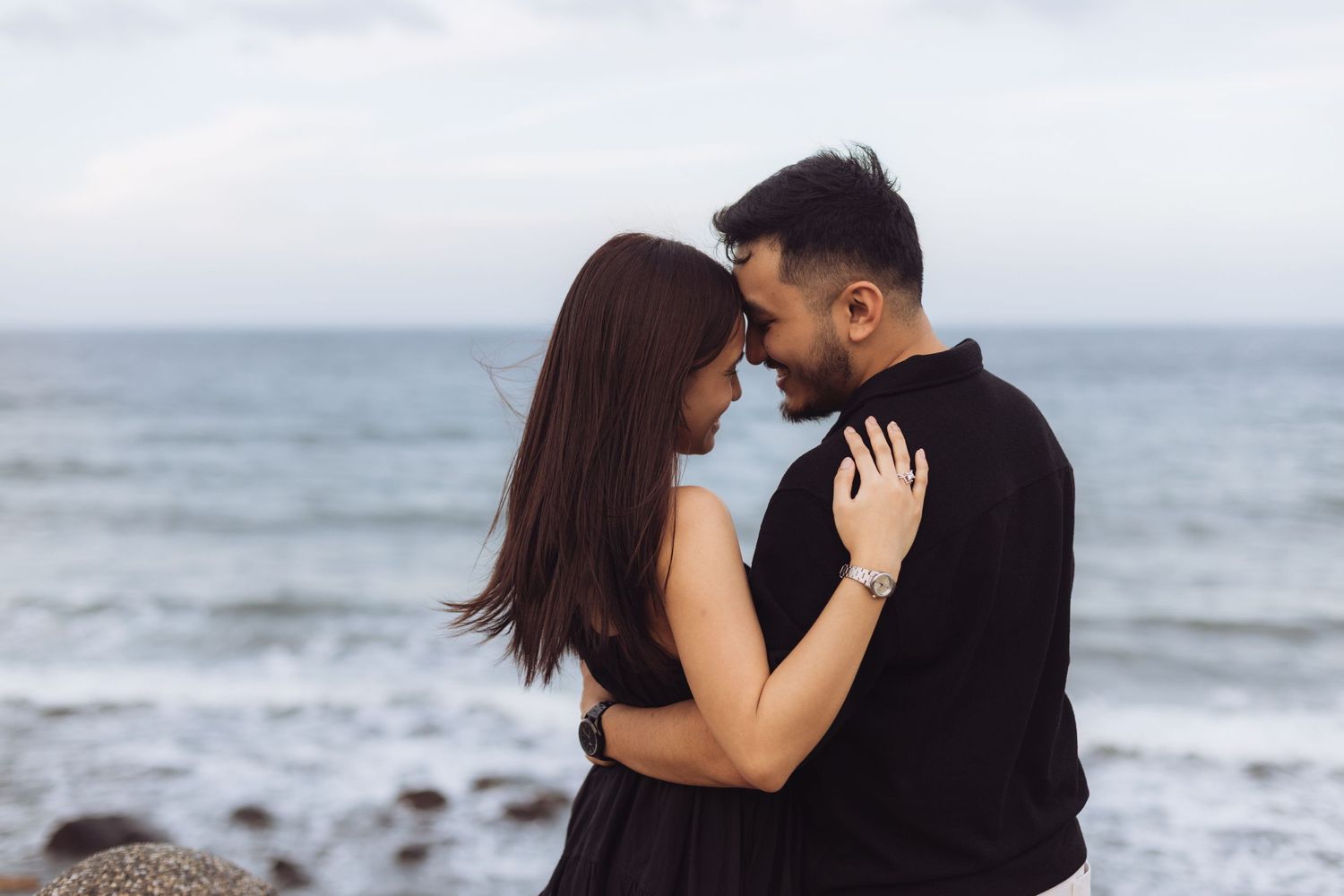 Romantic couple embracing on a rocky beach with ocean waves in the background on a cloudy day.