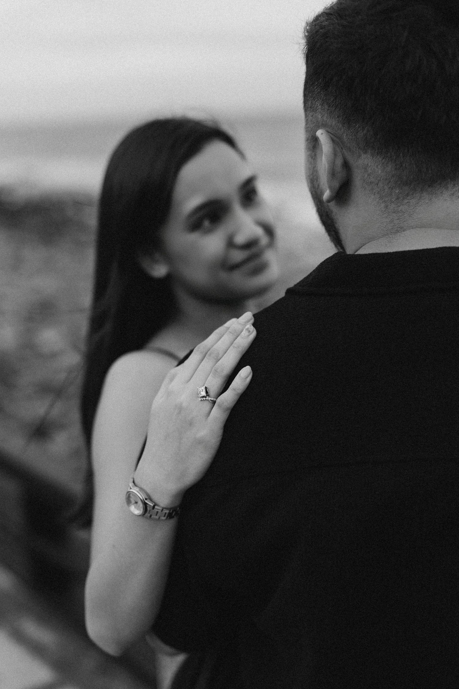 Black and white photo of a couple, woman placing hand on mans shoulder wearing a ring and watch.