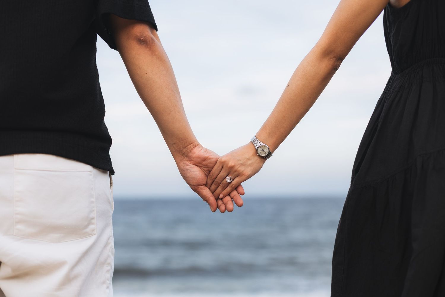 Couple holding hands at the beach, woman wearing a watch and ring, both dressed in black and white.