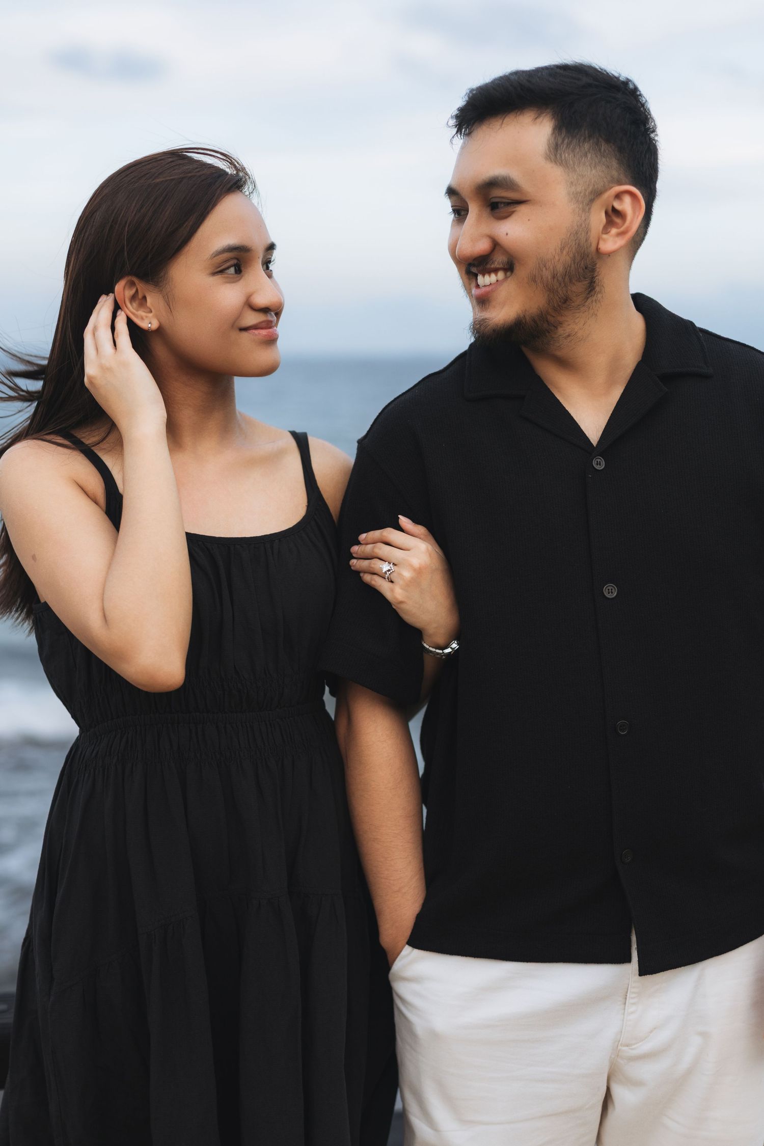 A smiling couple dressed in black poses together near the ocean, with the woman touching her hair.