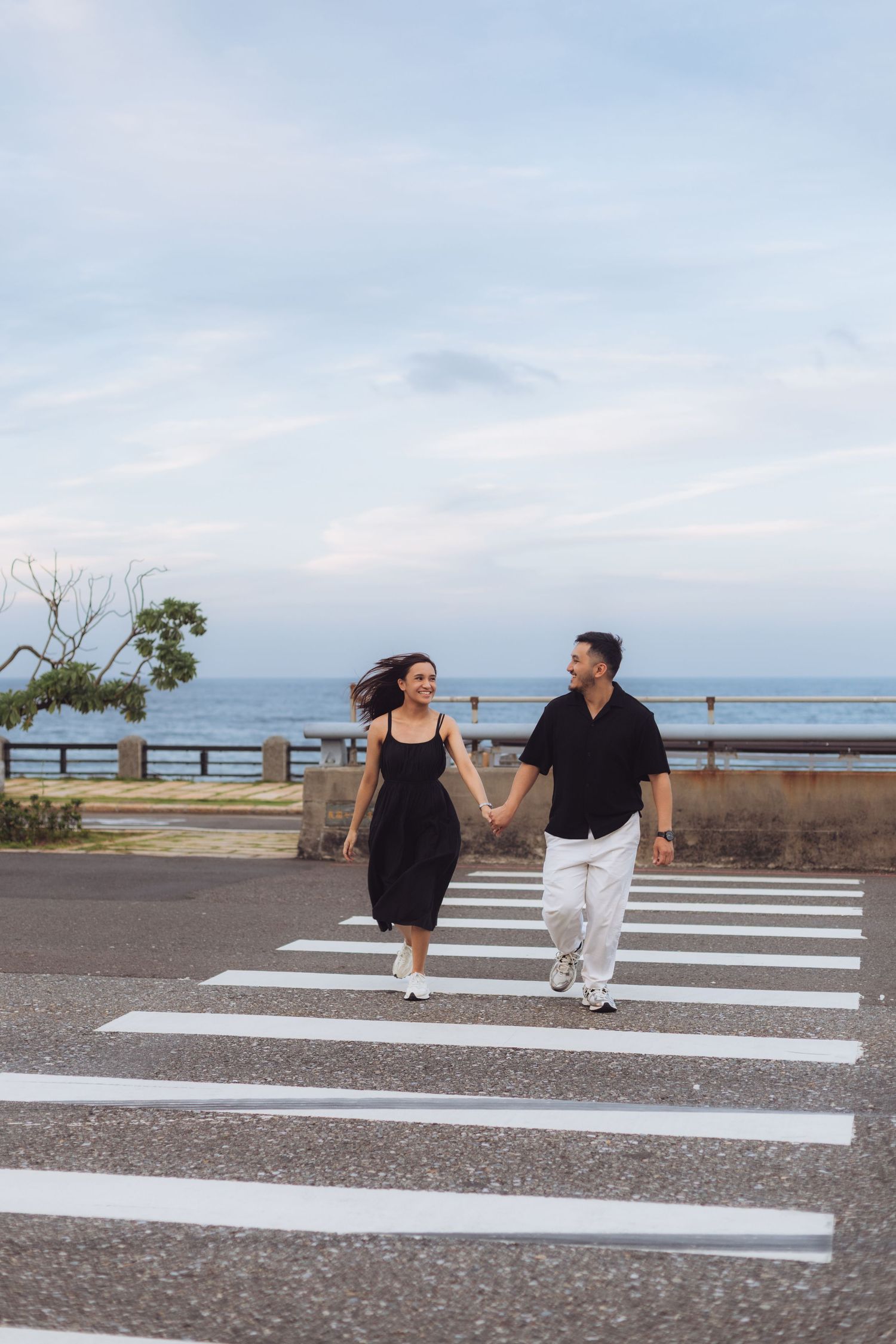 Couple holding hands and smiling while crossing a zebra crossing near the ocean on a cloudy day.