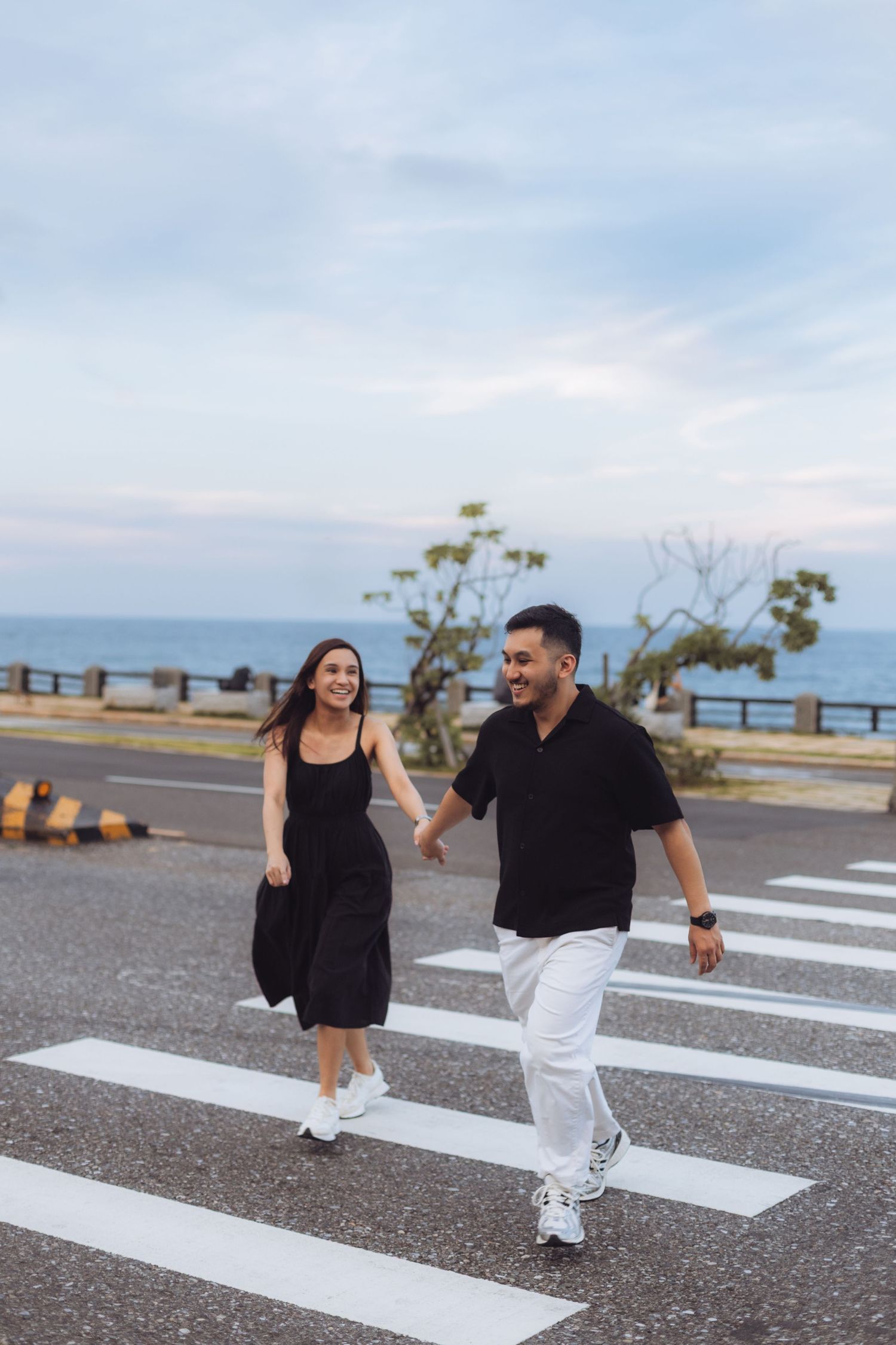Smiling couple holding hands crossing a zebra crosswalk near the ocean, wearing matching black outfits with white sneakers.