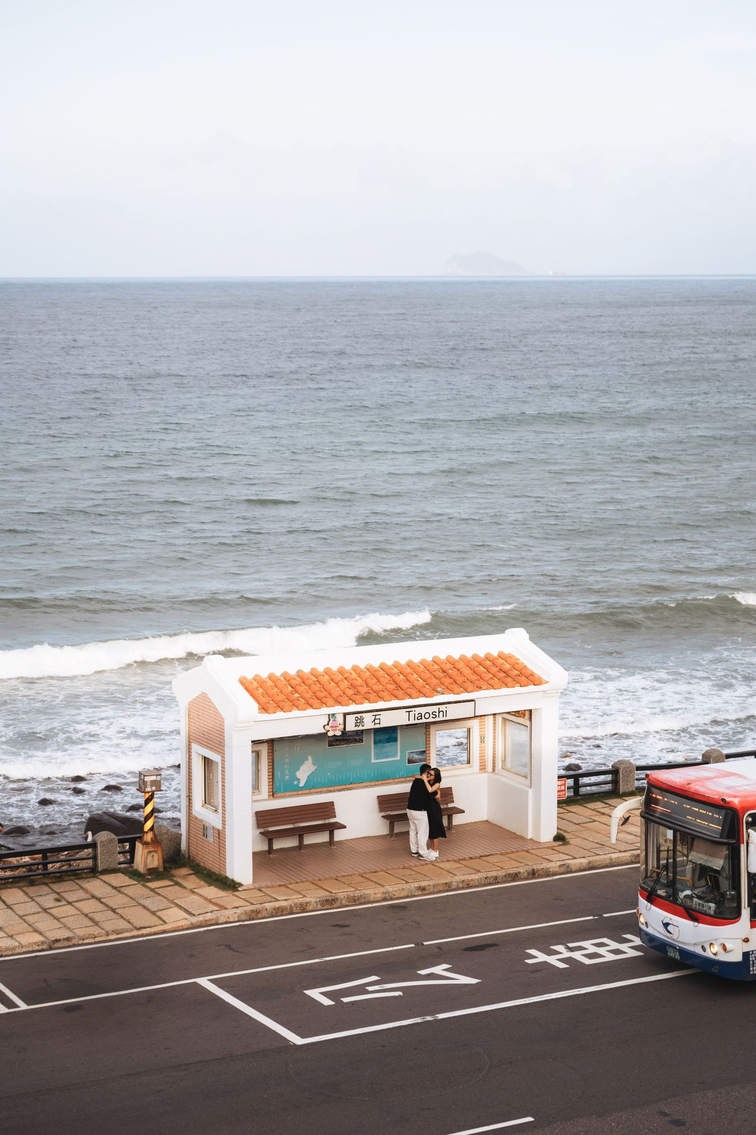 A scenic coastal bus stop with orange tile roof overlooks the ocean in Jeju, South Korea, with a couple waiting inside.