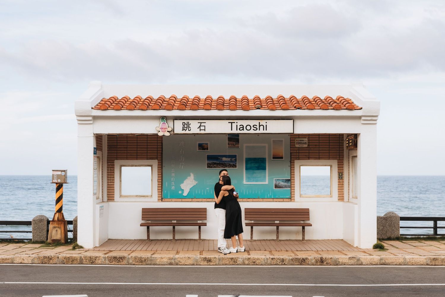 Couple embracing at Tiaoshi bus stop in Taiwan, with ocean views and red-tiled roof shelter on a cloudy day.