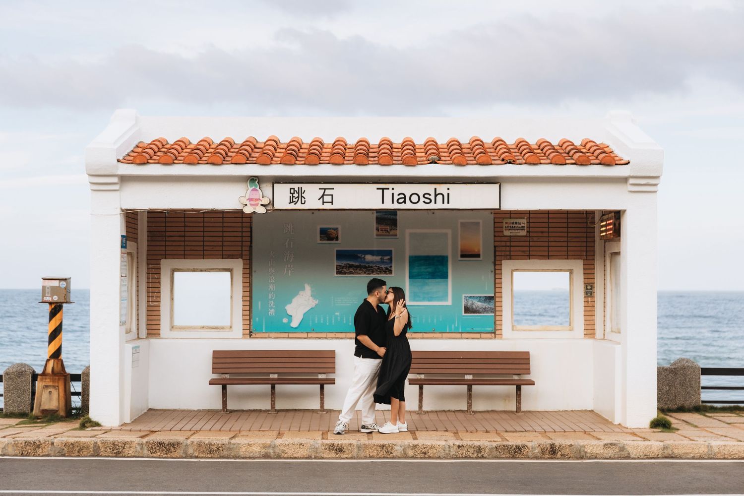 Couple embracing at Tiaoshi bus stop in Taiwan with ocean views and traditional red-tiled roof.