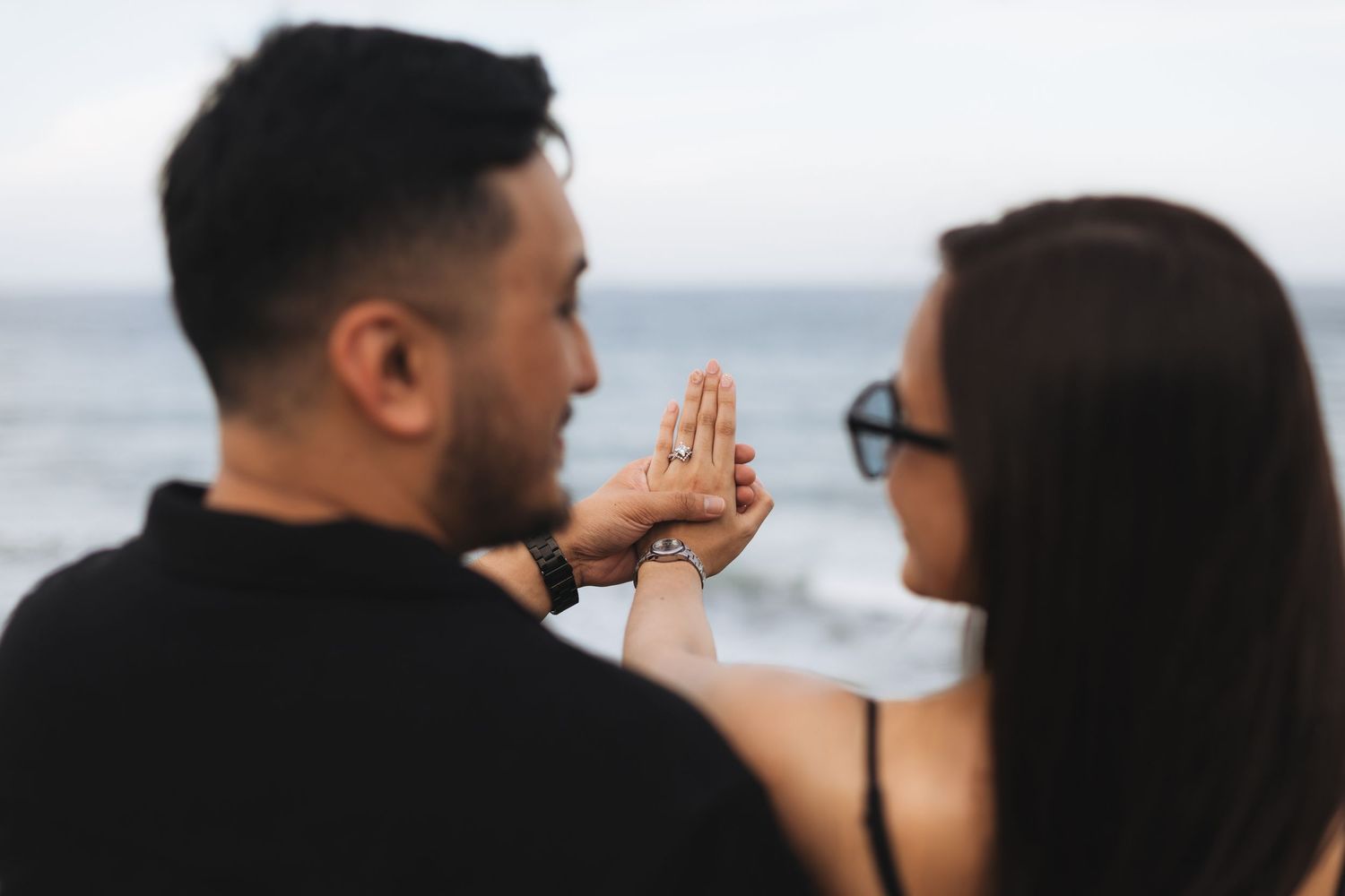 Couple holding hands near the ocean, woman showing engagement ring on beach.