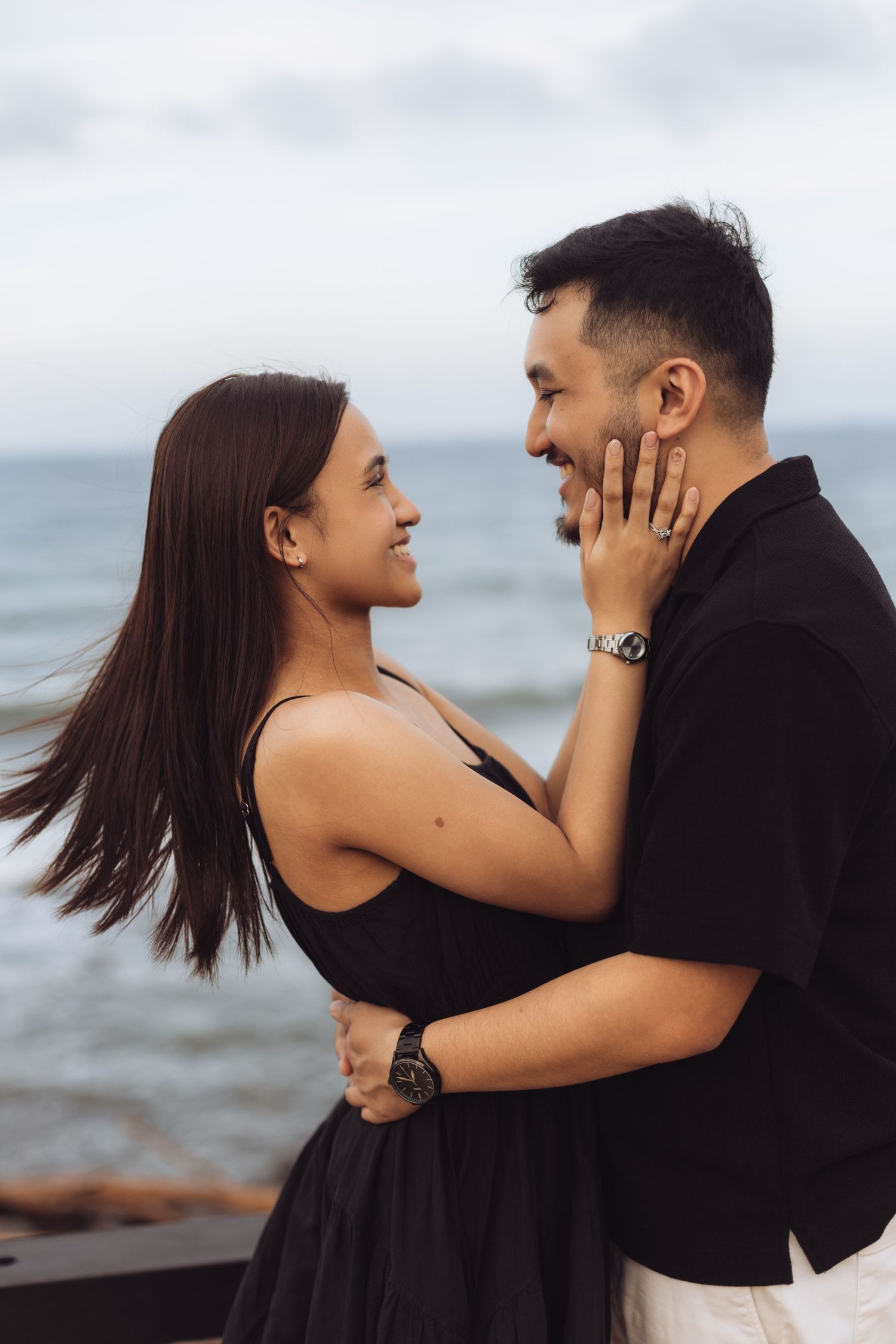 Couple in black outfits embracing and smiling at each other on a beach with ocean in the background.