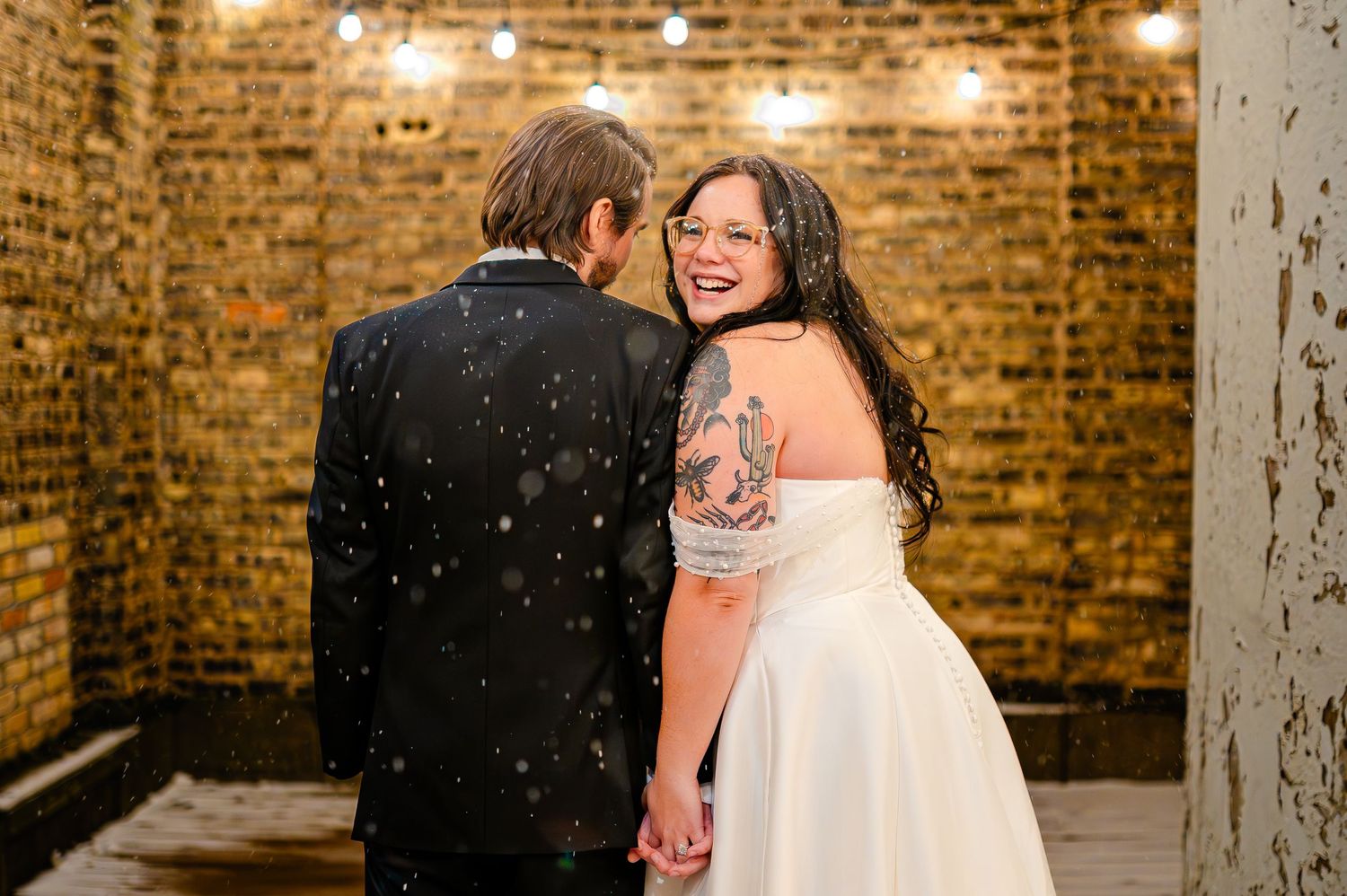 Bride with tattoos in white off-shoulder gown smiles back at camera while holding groom's hand in snowy brick venue.
