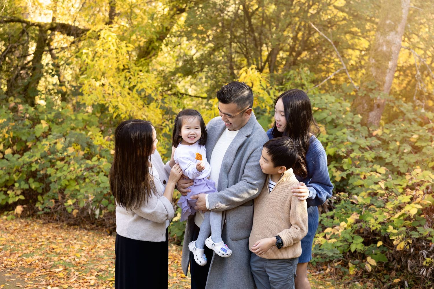 Family of five poses outdoors in autumn park with golden foliage, parents holding young children in fall setting.