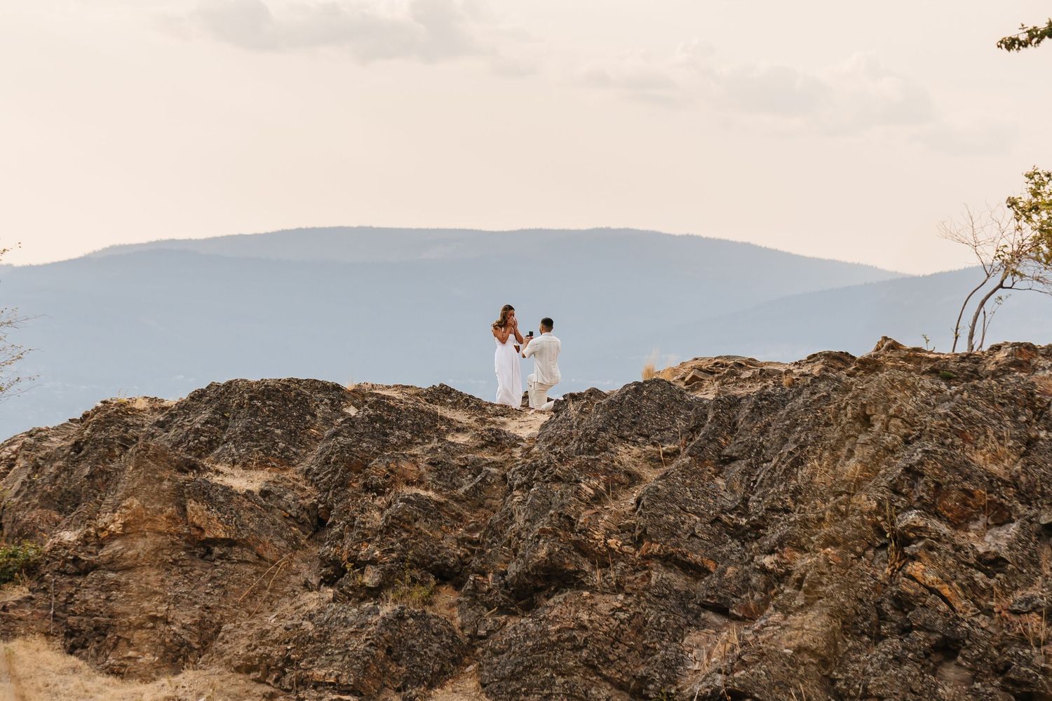 Couple in white attire standing on volcanic rocks with mountain views in the background.