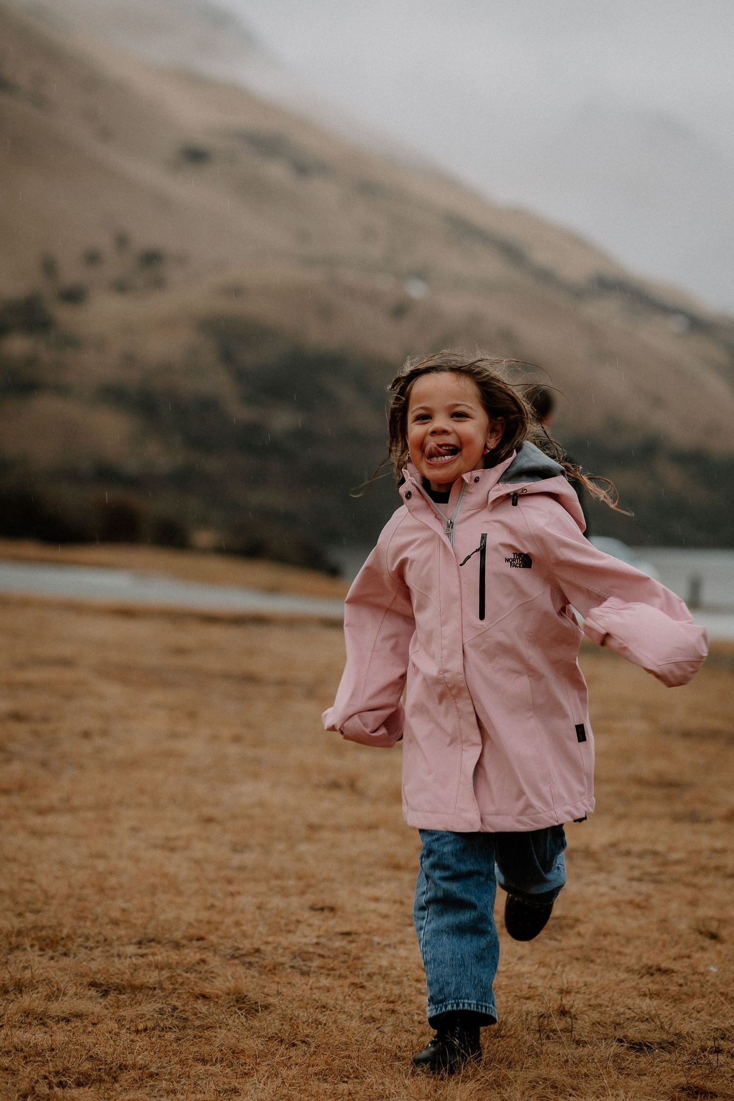 A joyful young girl in a pink jacket runs through a field with mountains and a lake in the background.