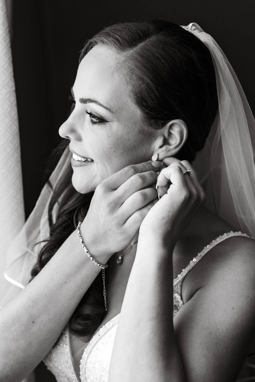 Black and white photo of a bride putting on earrings while wearing a veil and wedding dress on her wedding day.
