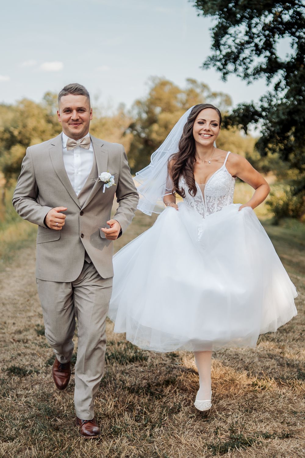 Happy bride and groom walking outdoors in nature, bride in short white tulle dress with veil, groom in beige suit.