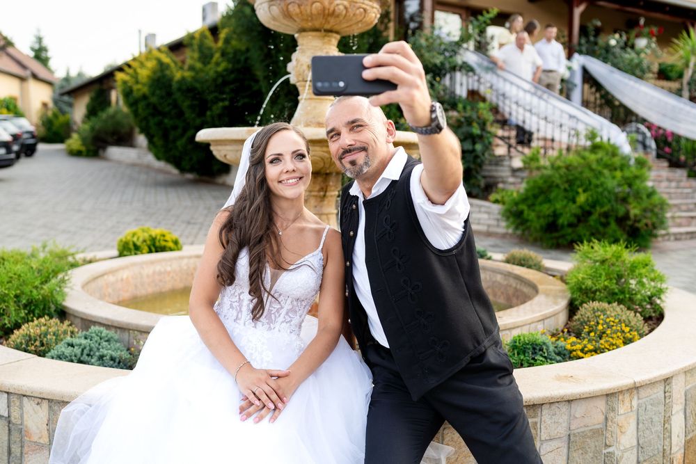 Bride and groom taking a selfie together near a stone fountain outdoors on their wedding day.
