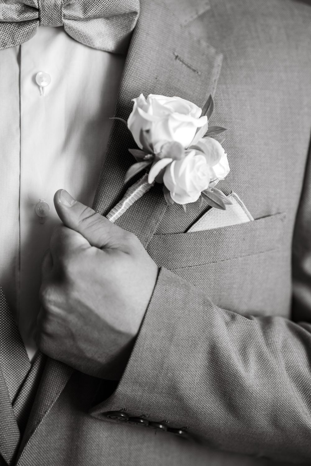 Black and white photo of a groom adjusting his boutonniere with white roses on a suit lapel.