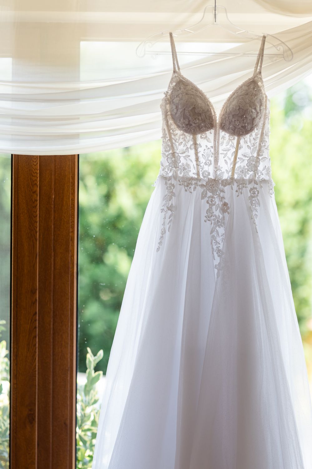 Elegant A-line wedding dress with lace bodice and tulle skirt hanging by a sunlit window.