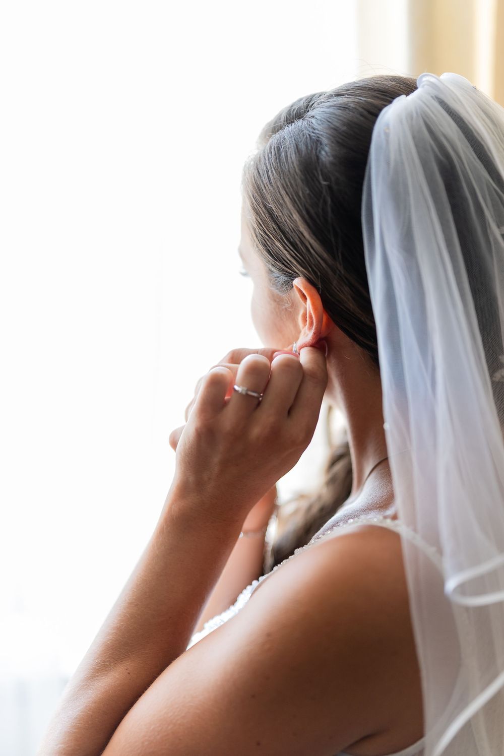 Bride in white veil putting on earring on her wedding day, viewed from behind near a bright window.