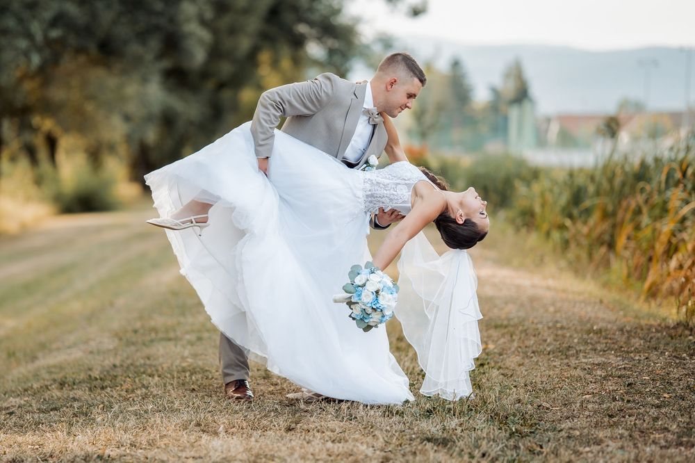A groom dips his bride in a romantic outdoor wedding dance pose, her white gown flowing as she holds a blue bouquet.