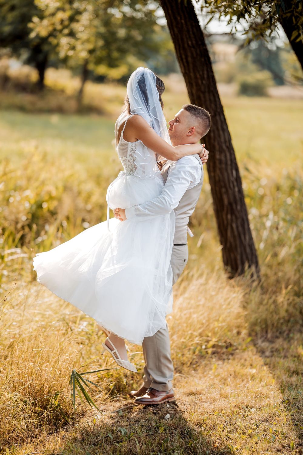Groom lifting bride in white dress and veil outdoors in sunny field near trees on wedding day.