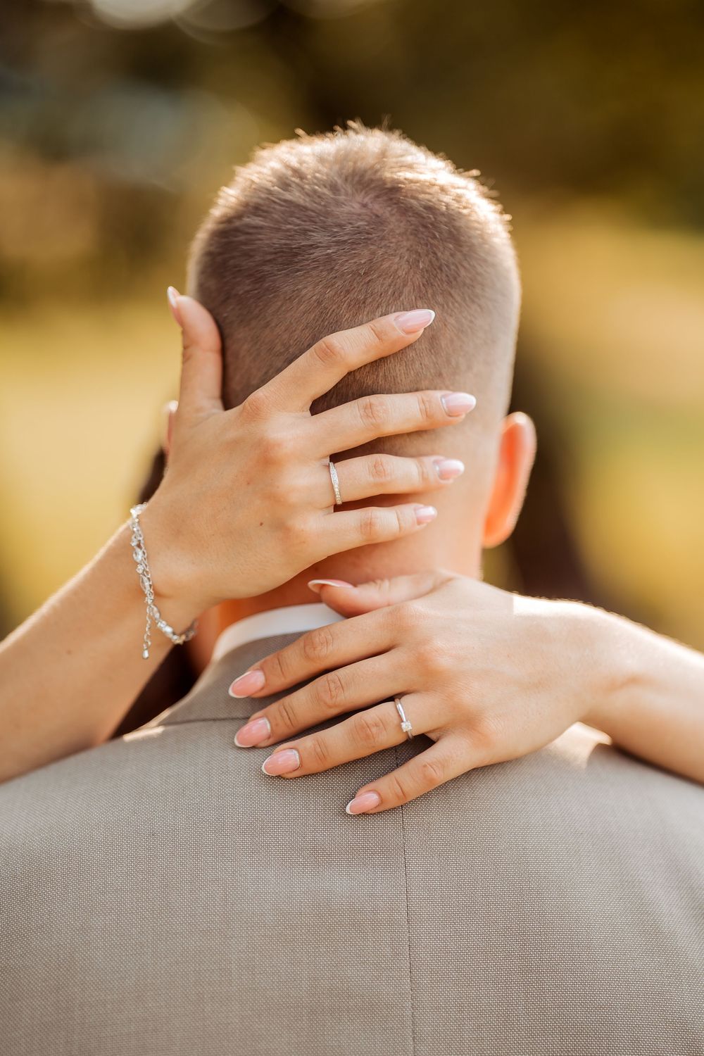 Woman with engagement ring and bracelet embracing man from behind, outdoor golden hour bokeh background.