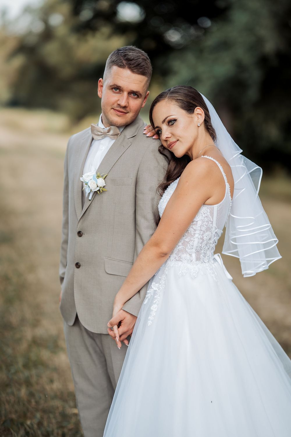 A bride in a white lace gown and veil holds hands with a groom in a gray suit outdoors.