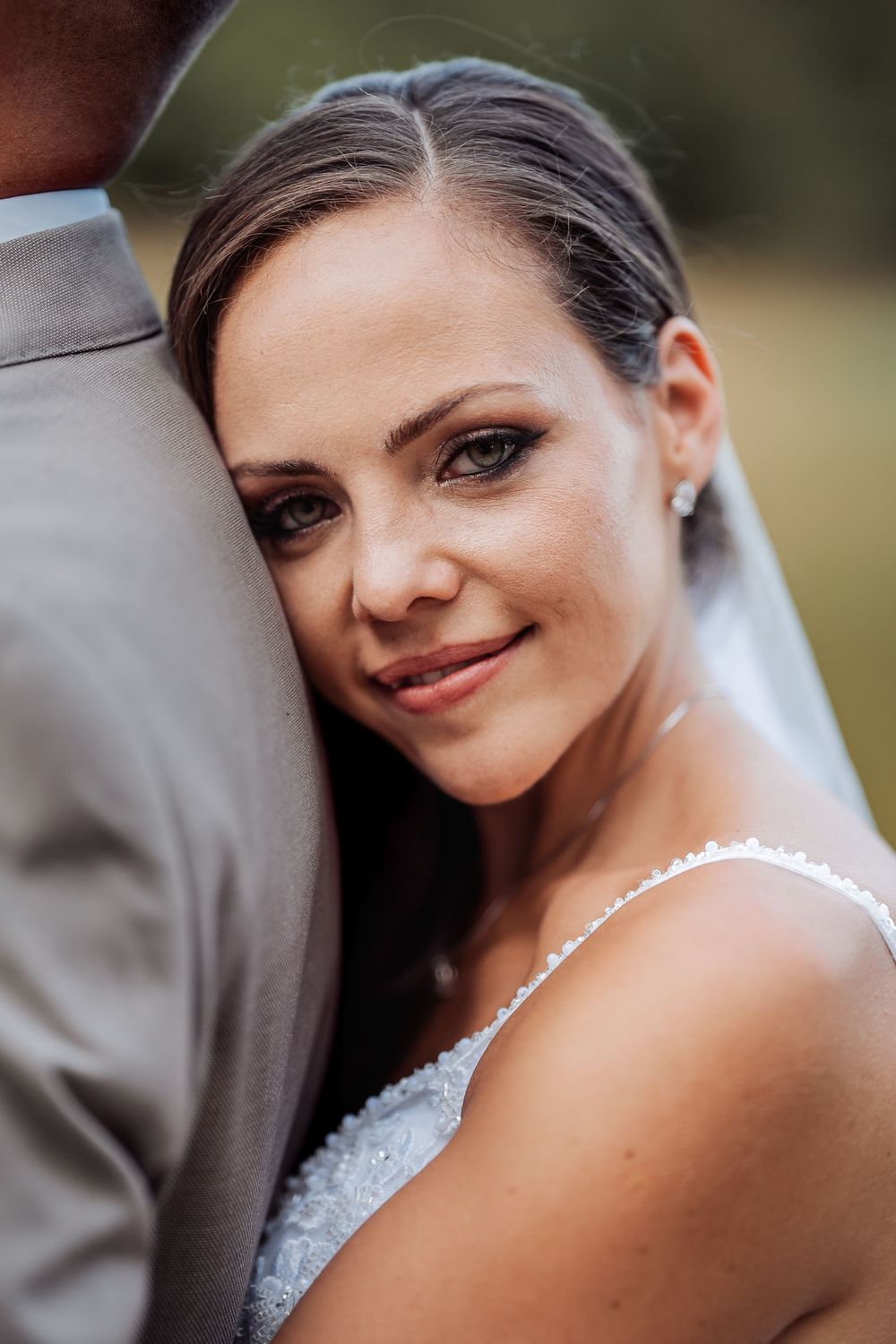 Bride in white lace dress leans against groom in gray suit, smiling softly at camera on wedding day.
