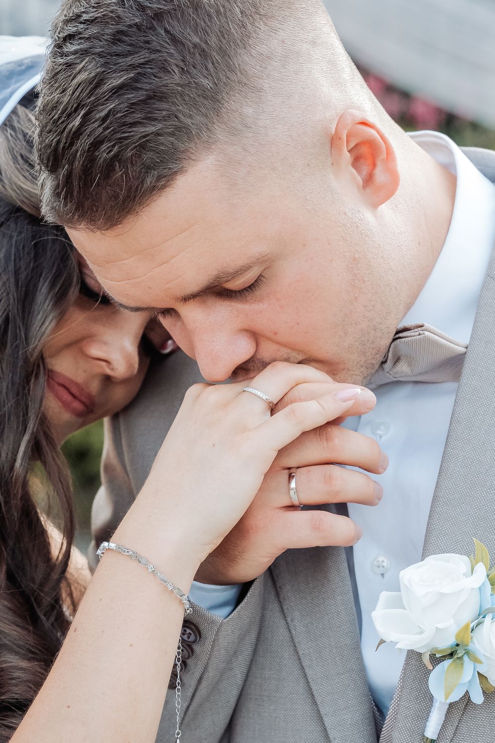 Groom in gray suit kisses bride's hand tenderly, both wearing wedding rings, with white boutonniere visible.