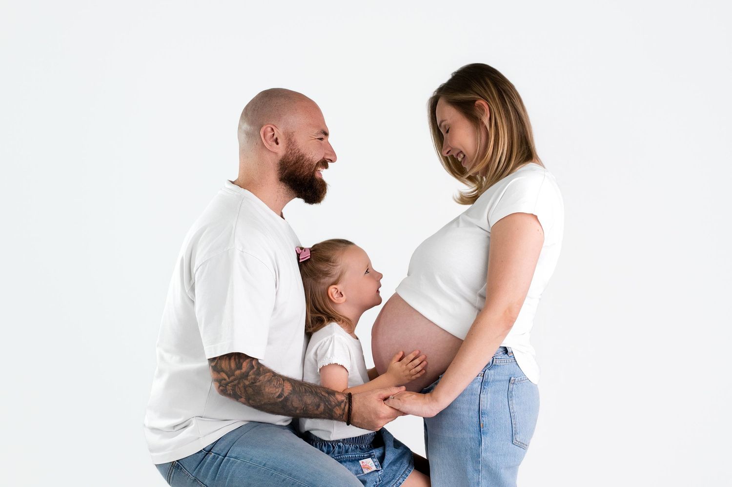 Pregnant woman and bald tattooed man smile at toddler child between them in white studio maternity photo.