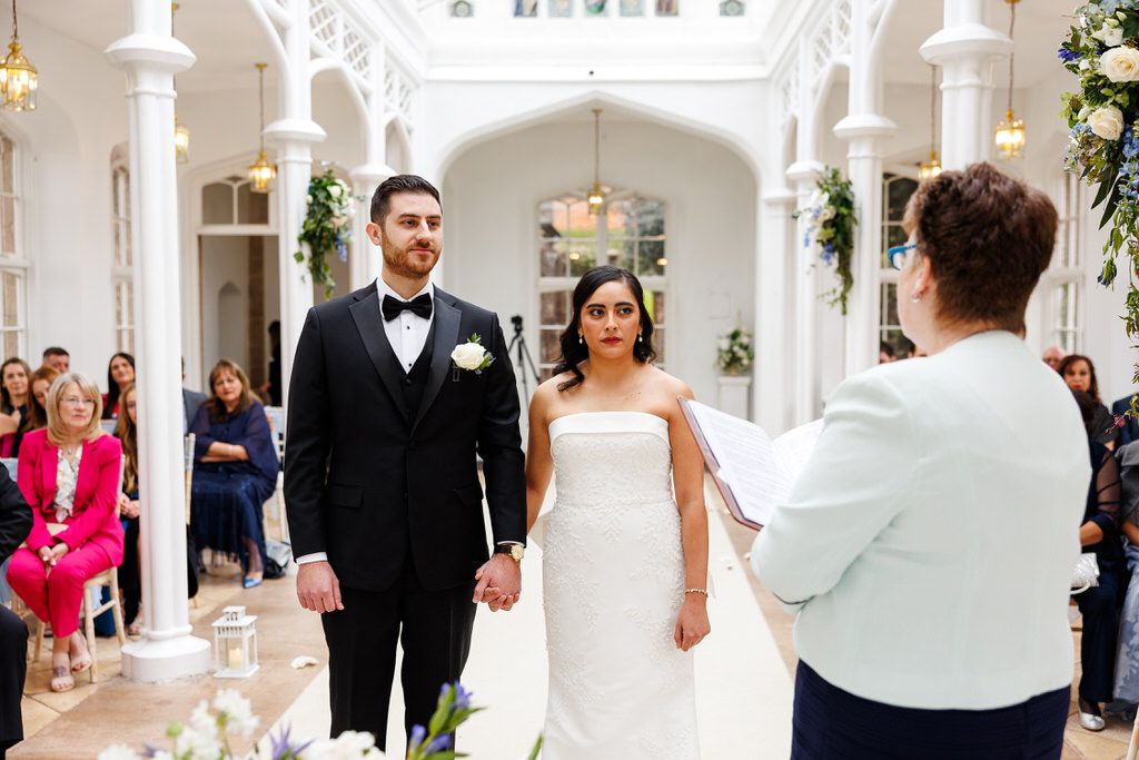 Bride and groom hold hands at elegant wedding ceremony in bright white venue with floral arch decorations.