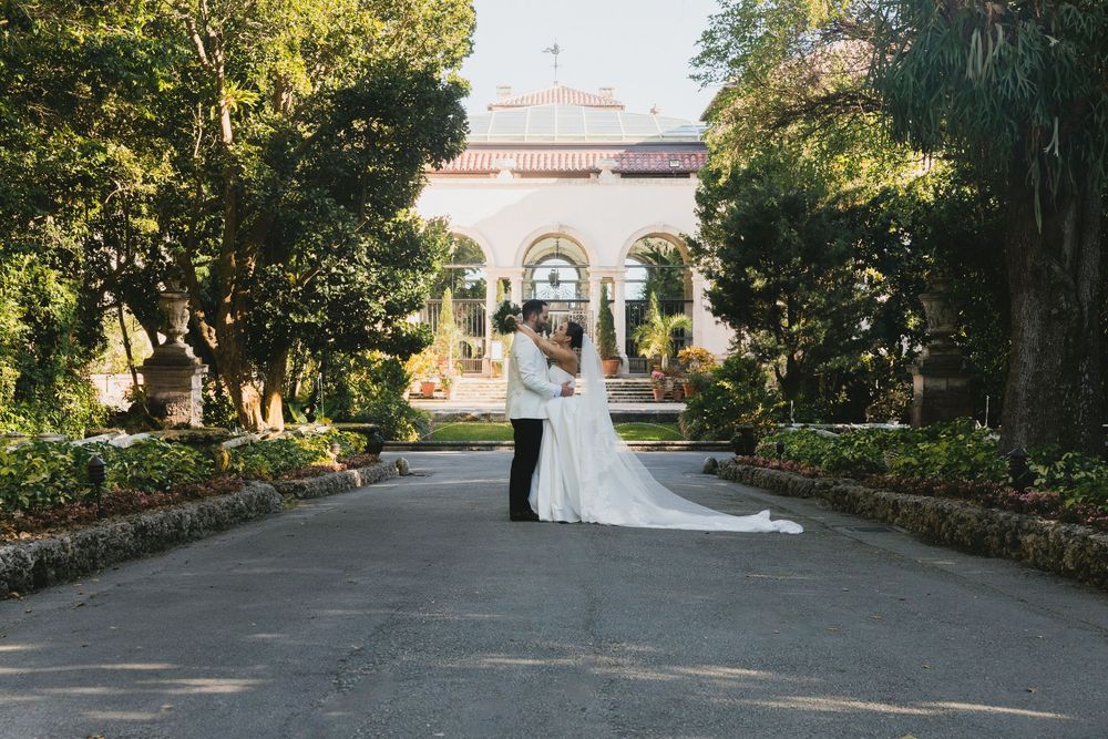 Bride and groom share a romantic kiss on a tree-lined path leading to an elegant white wedding venue.