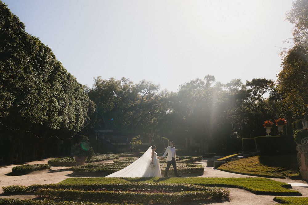 Bride and groom walk hand in hand through a sunlit formal garden with manicured hedges and tall trees.