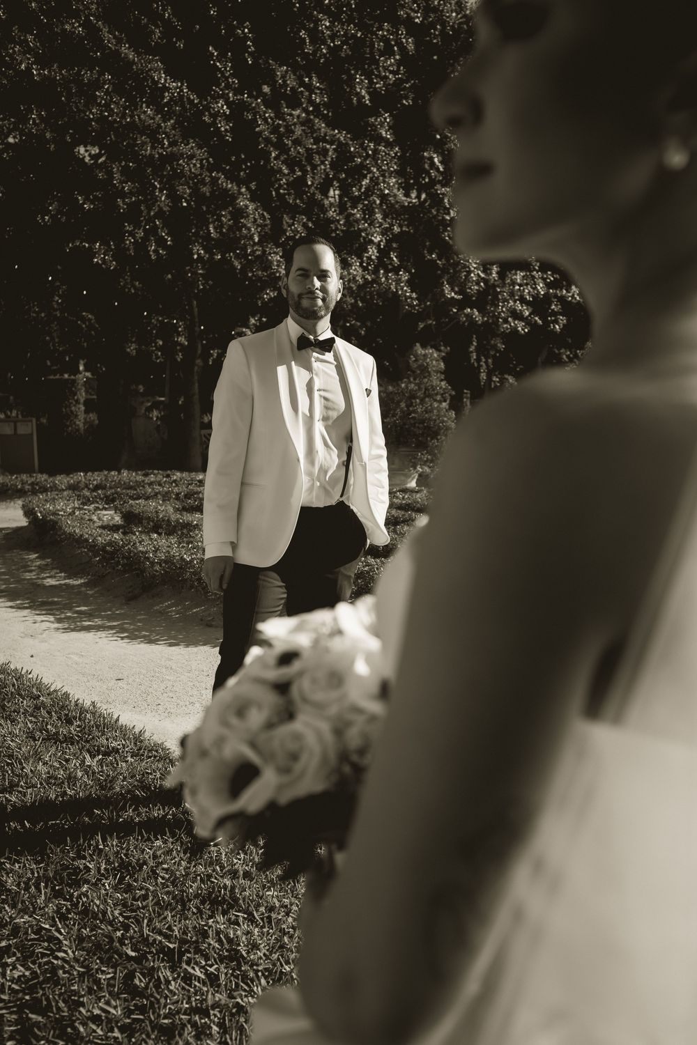 Black and white wedding photo of groom in white tuxedo watching bride holding bouquet in garden.