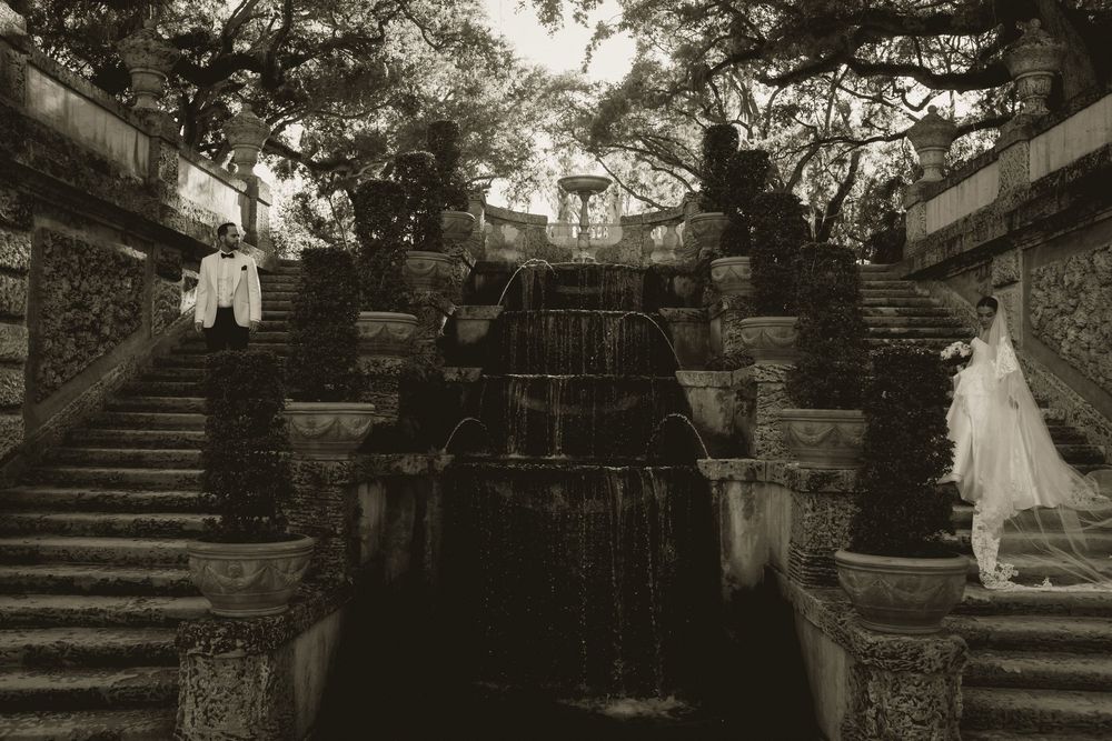 Black and white wedding photo of bride and groom at ornate stone fountain with cascading waterfall and grand staircase.