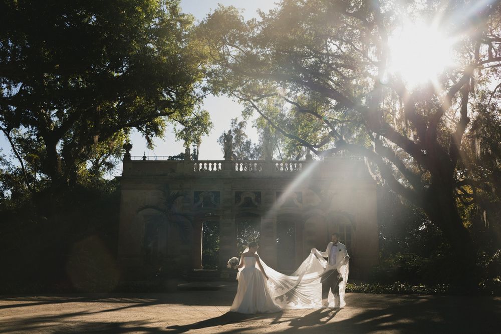 Bride and groom stand in golden sunlight before a grand estate, her veil flowing dramatically in the breeze.