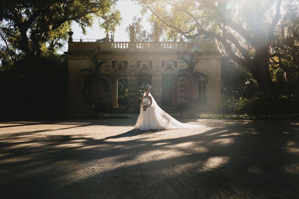 Bride and groom embrace in golden sunlight before an elegant stone mansion with balustrade, long veil trailing on cobblestone.