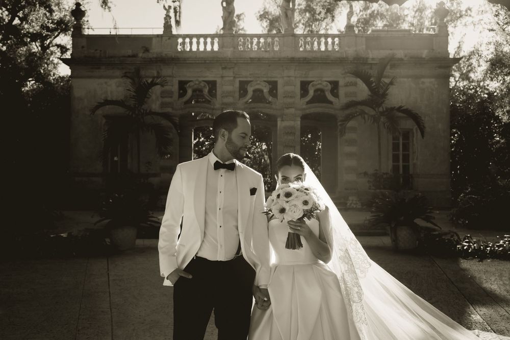 Black and white photo of a bride and groom standing together outside an elegant mansion on their wedding day.