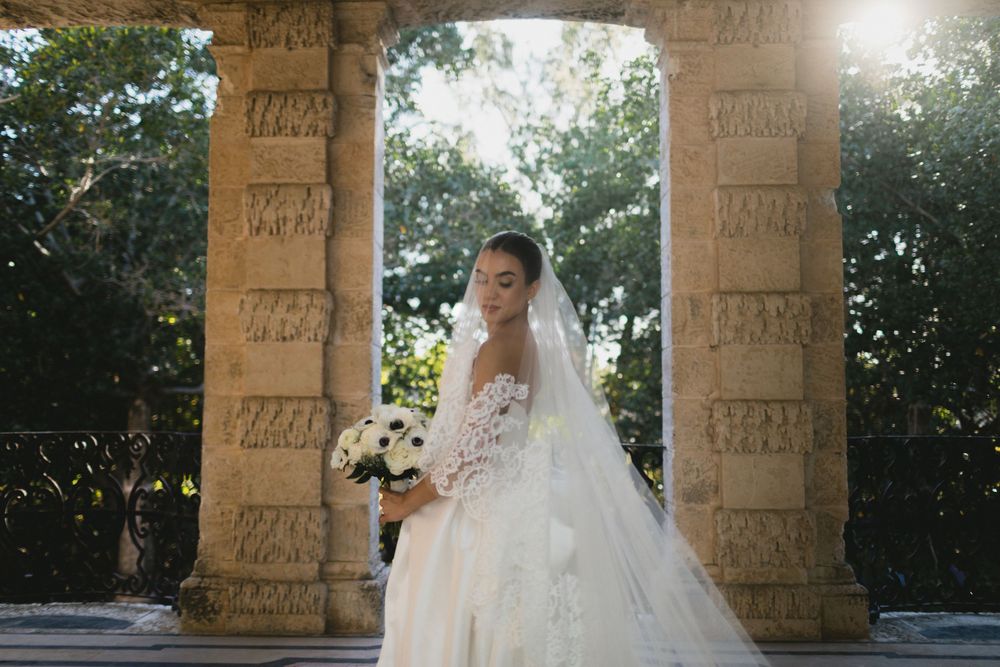 Bride in elegant lace wedding gown holding bouquet under ornate stone archway surrounded by lush greenery.