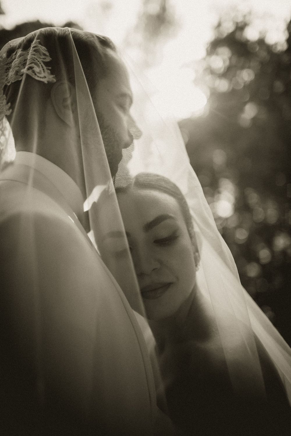 Black and white wedding photo of bride and groom sharing intimate moment under bridal veil with sunlit bokeh background.
