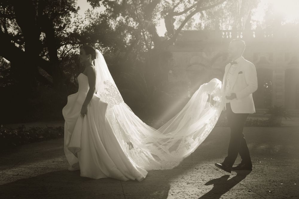 Black and white wedding photo of bride in flowing veil and groom in white jacket walking outdoors in sunlight.