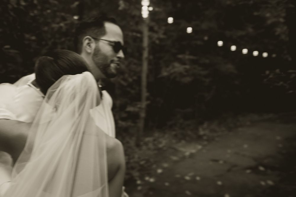 Groom carrying bride in a black and white photo with string lights glowing in a wooded setting.